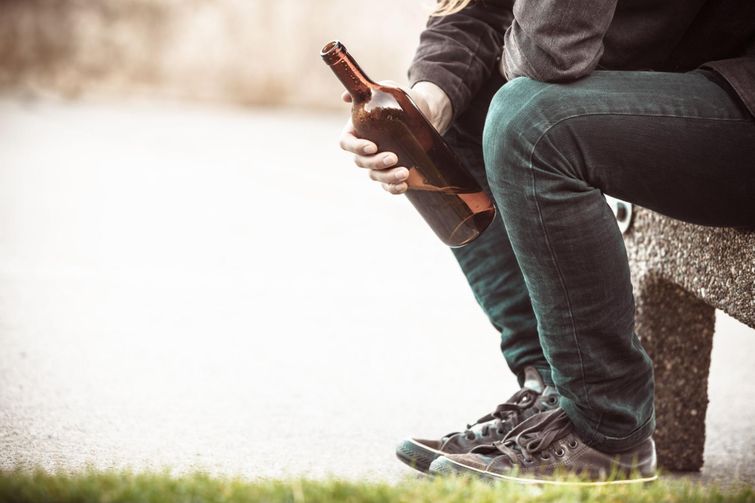 Young man with alcohol addiction holding beer bottle on concrete bench